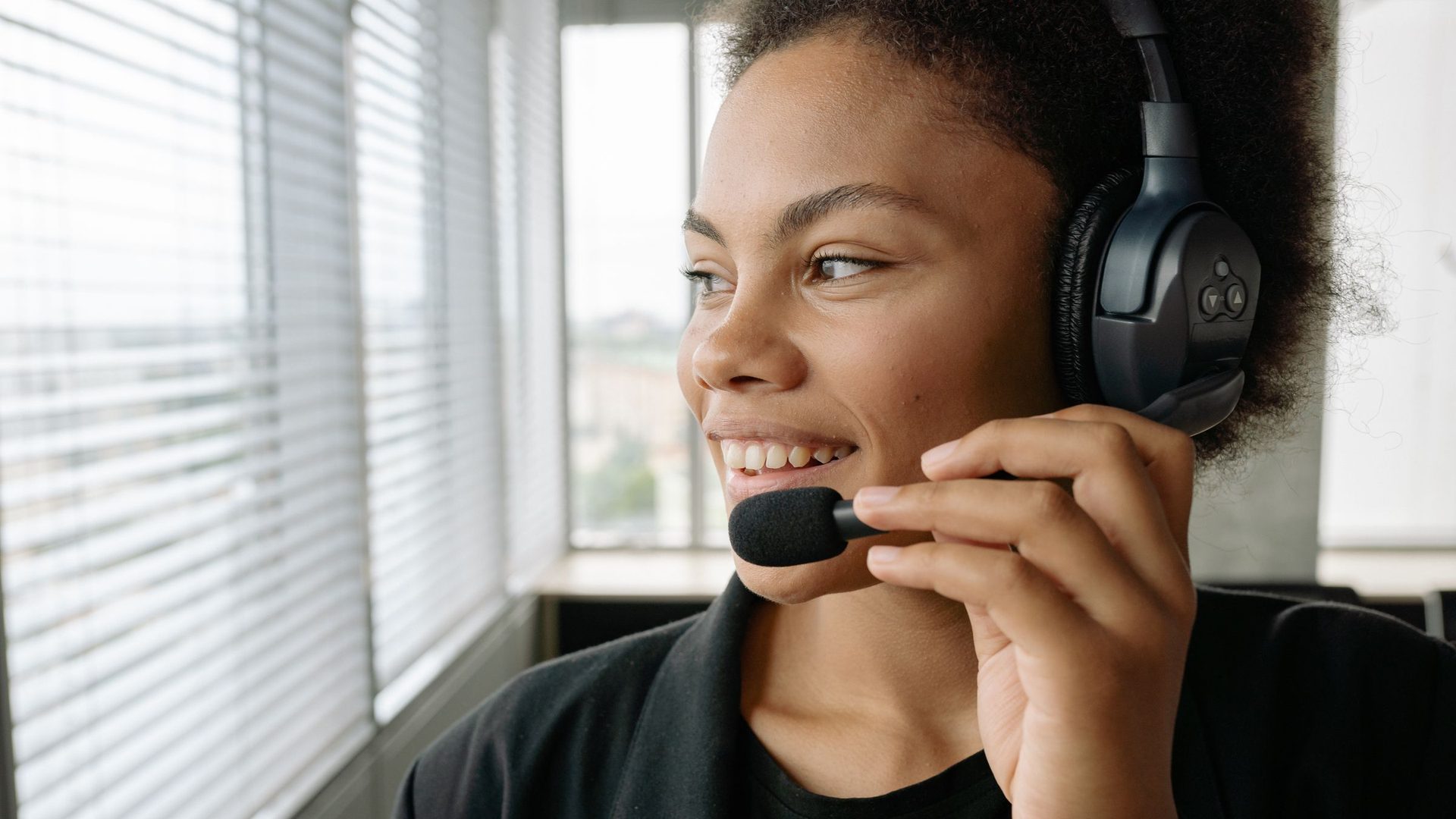 Une conseillère du service client sourit, casque et micro sur la tête, près d'une fenêtre.