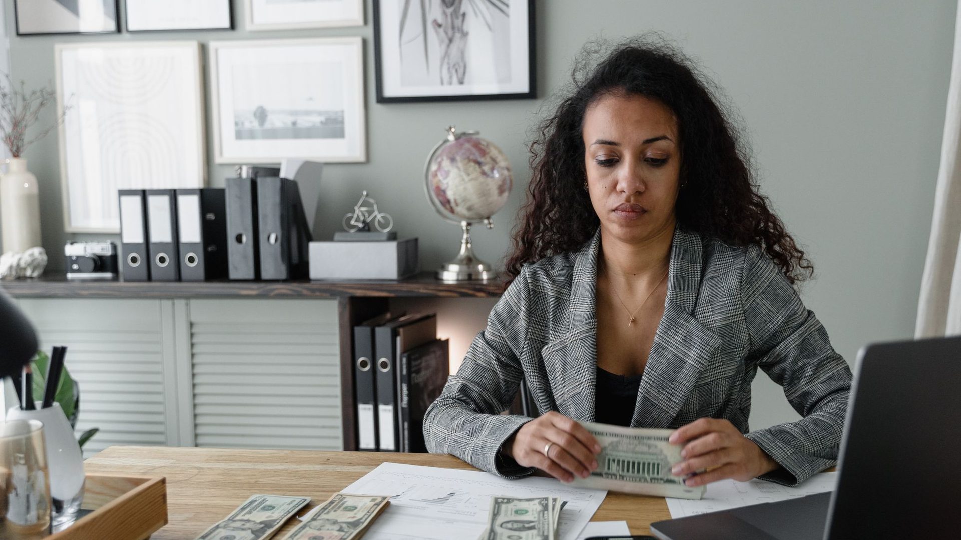 Une femme en veste compte des billets à son bureau, calculatrice et ordinateur à côté.