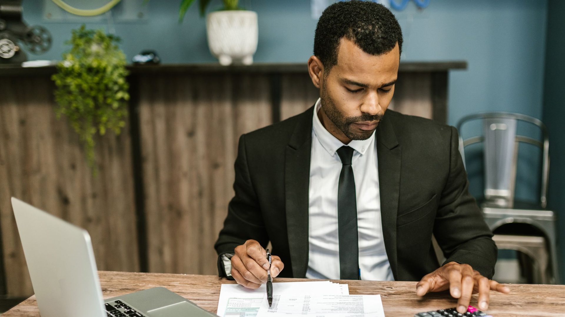 Un comptable en costume vérifie des factures à son bureau, stylo et calculatrice en main, l'ordinateur ouvert à côté.