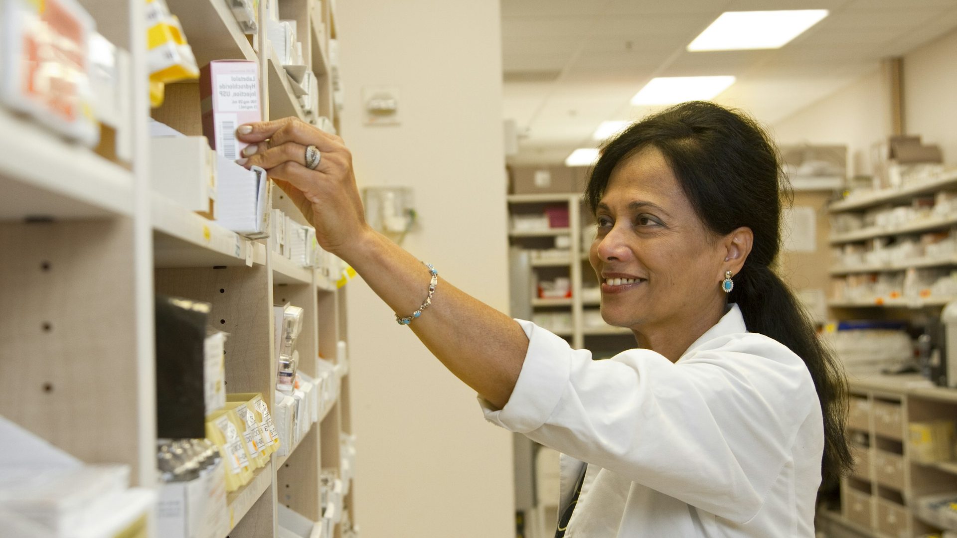 Une pharmacienne en blouse blanche prend une boîte de médicament sur une étagère de la réserve.