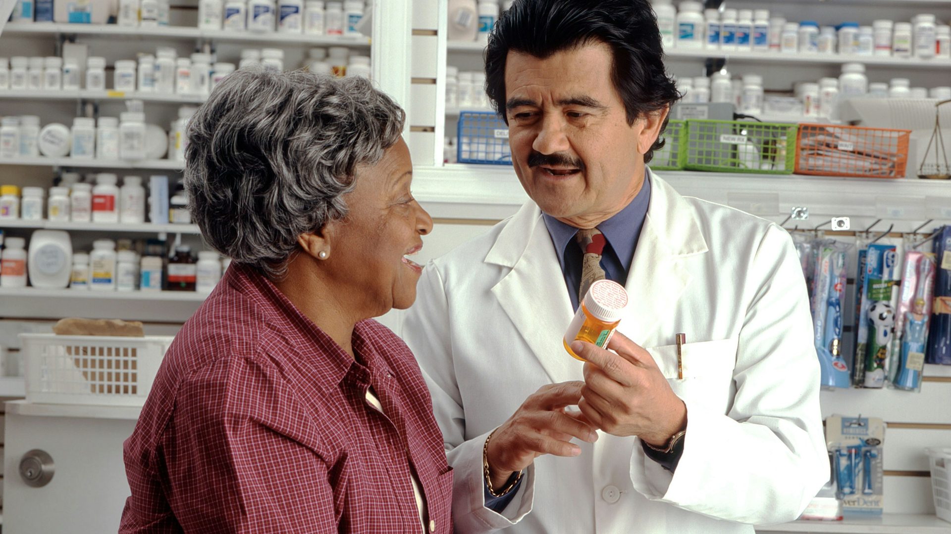 Un pharmacien en blouse montre un flacon de médicament à une cliente, au comptoir de son officine.